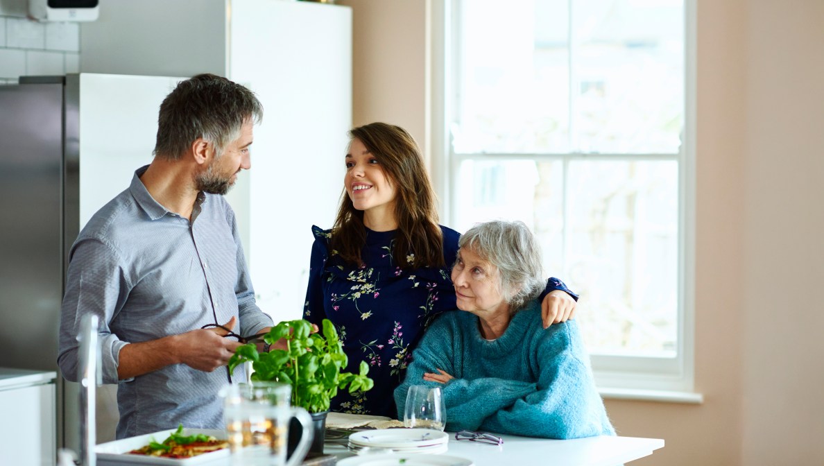 young woman standing in the kitchen with her arm around an older woman and a man standing and smiling