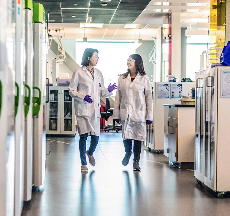 Two lab technicians in white lab coats having a conversation while walking in lab
