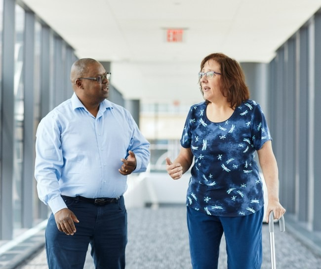 Woman walking with cane walks down hallway talking with man
