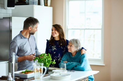 young woman standing in the kitchen with her arm around an older woman and a man standing and smiling